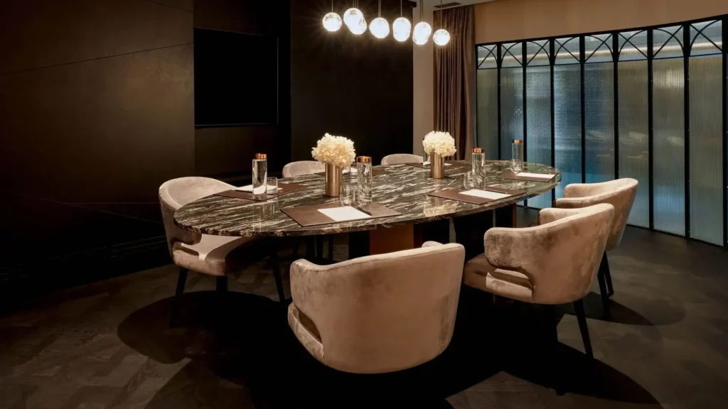 Modern conference room with an oval marble table, eight beige chairs, bottled water, notepads, and decorative white flowers, lit by a row of globe pendant lights.
